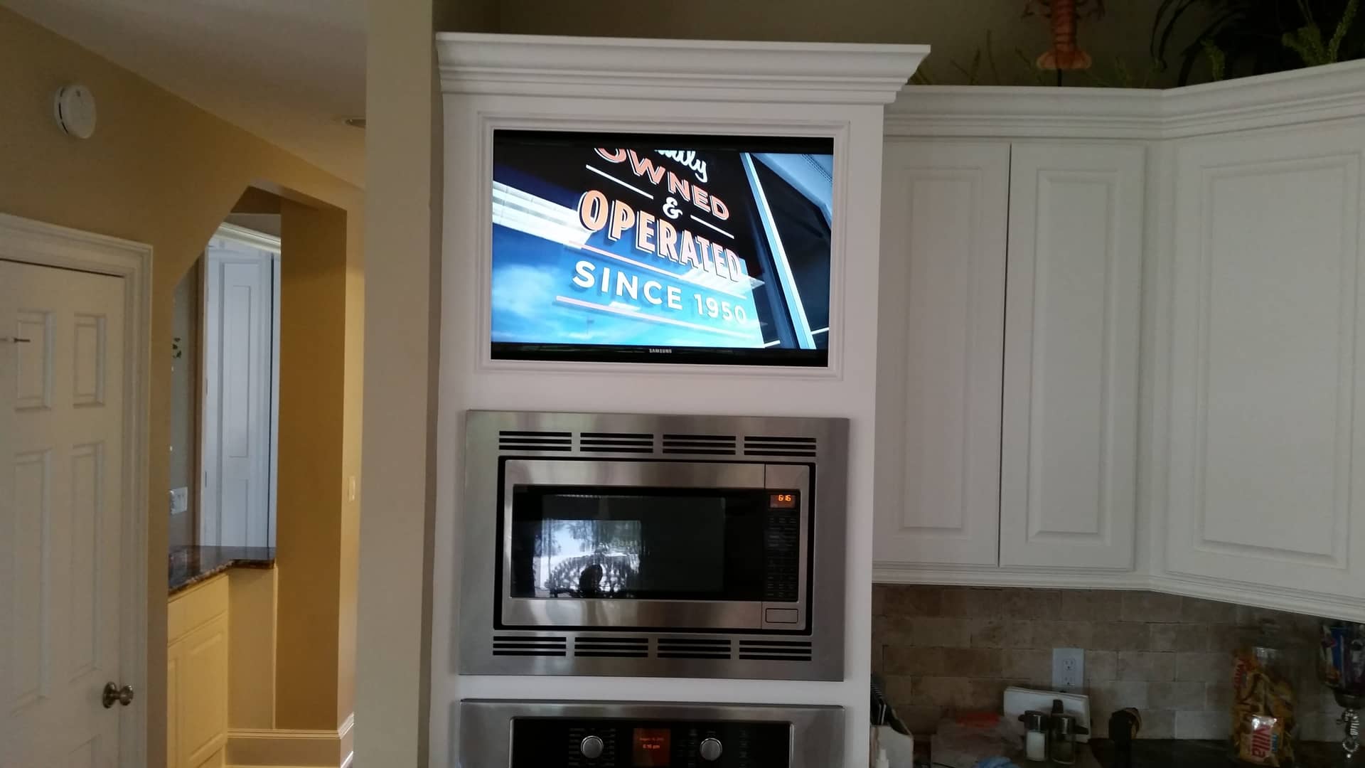 A modern kitchen setup with a flat-screen TV mounted above a built-in microwave, surrounded by white cabinetry.