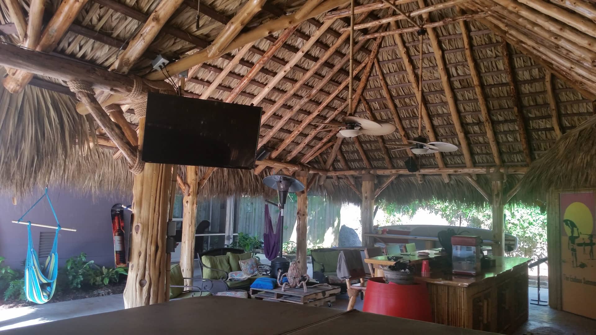 Outdoor beach bar under a thatched roof with a TV mounted on a wooden beam, surrounded by tropical decor.