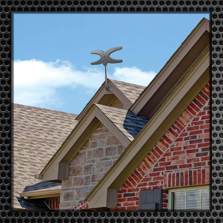 A close-up view of a metallic antenna mounted on the peak of a residential roof with brick details and shingles.