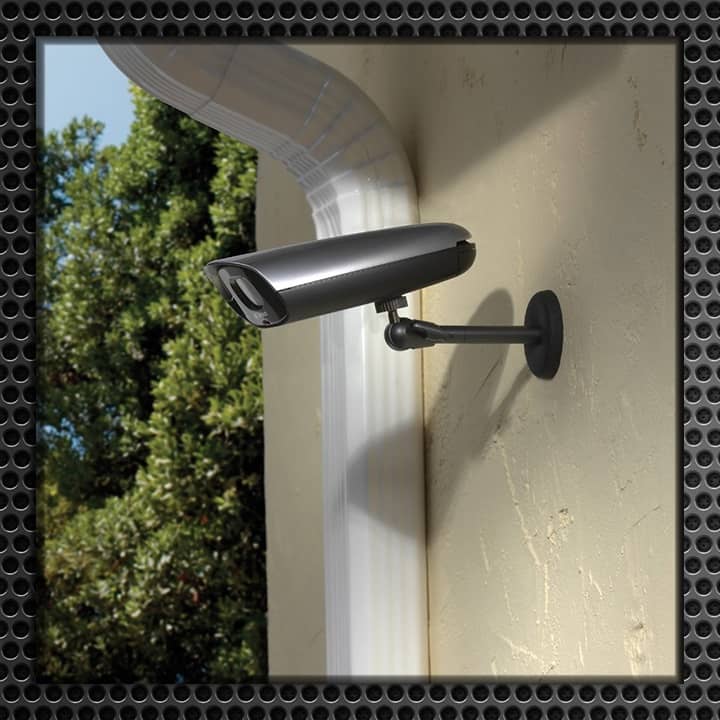 A modern security camera mounted on a light-colored exterior wall, overlooking a garden area.