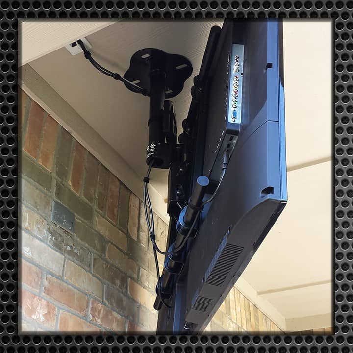 Close-up of a ceiling mount holding a flat-screen TV under an outdoor patio cover, showing detailed mount mechanism and cable management against a brick wall.